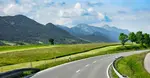 Curving country road in southern Germany with lush green fields and the Bavarian Alps in the distance under a partly cloudy sky — ideal scenic drive setting for a German road trip playlist.