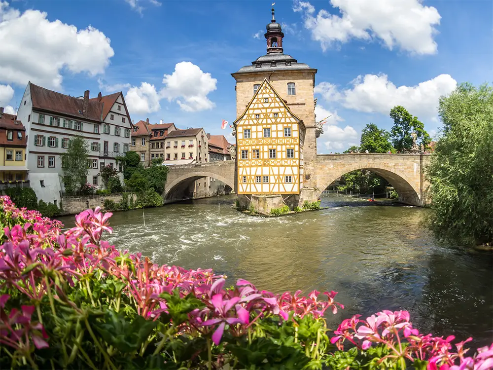 Historic town hall of Bamberg built over the Regnitz River, framed by blooming pink flowers.