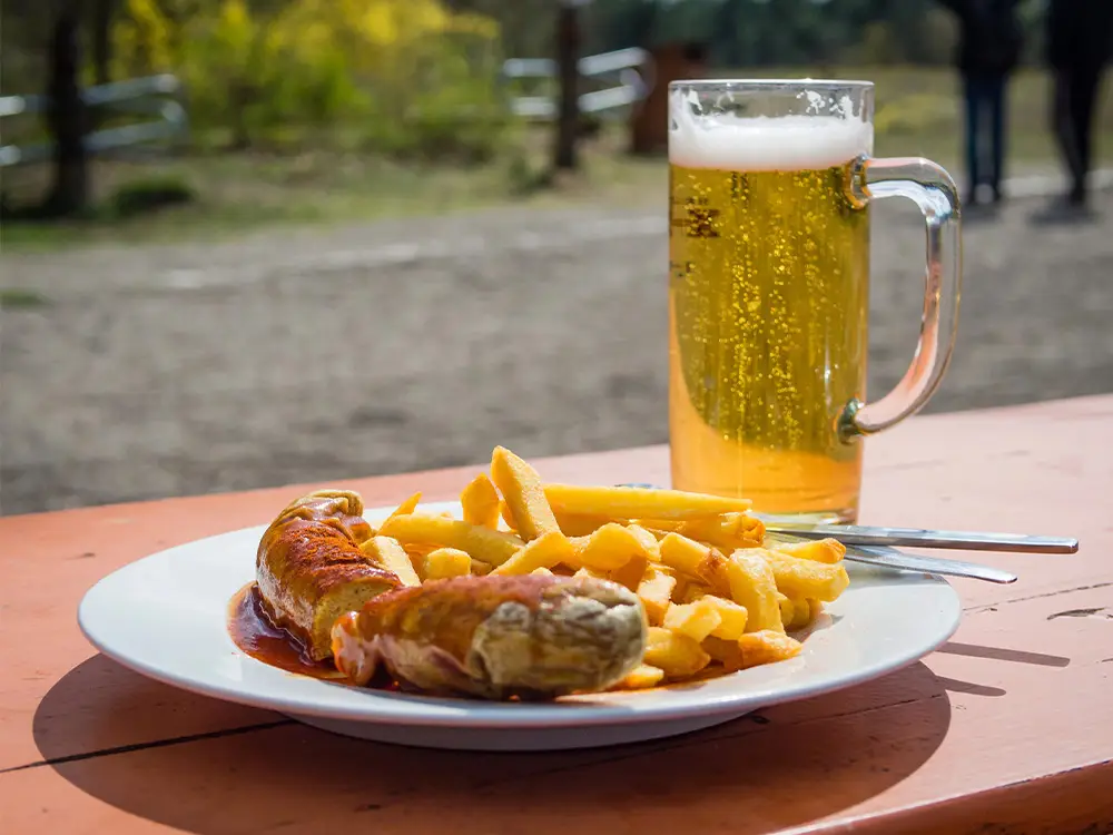 Traditional German currywurst with fries and a cold beer on an outdoor picnic table.