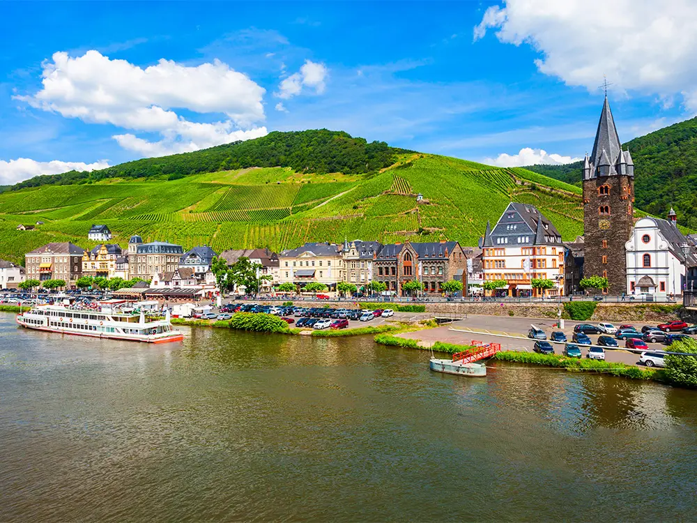Riverside view of Bernkastel-Kues with vineyards in the background and a river cruise boat docked.