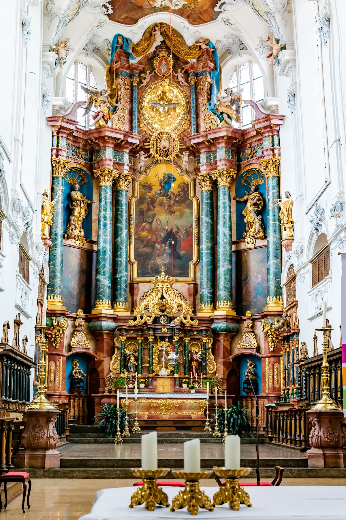 Intricate baroque altar with ornate gold detailing, statues, and a large painting, surrounded by white walls and high ceilings. Candles on a foreground table.