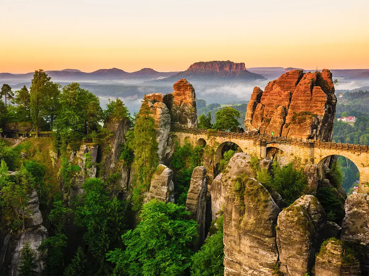 A stone bridge spans between tall, rocky formations surrounded by lush greenery at sunrise. Mist lingers in the distant valley, with hills silhouetted against the sky.