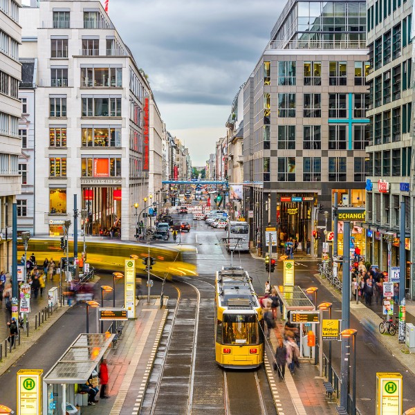 View of Friedrichstrasse A busy urban street in Berlin, Germany, with trams, cars, pedestrians, and tall buildings on both sides. Traffic lights, street signs, and tram platforms are visible. Cloudy sky overhead.