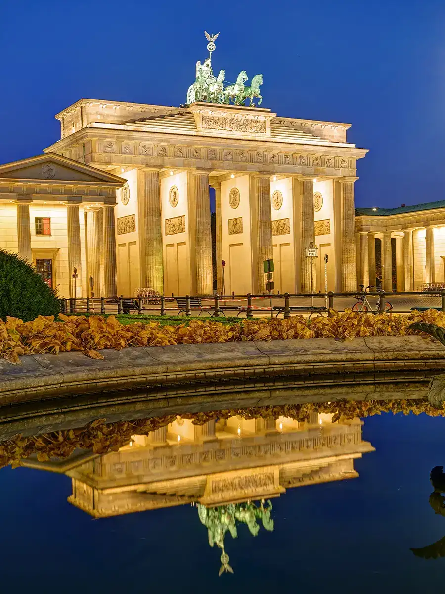 The Brandenburg Gate in Berlin is illuminated at night, with its reflection visible in the water of a nearby fountain.