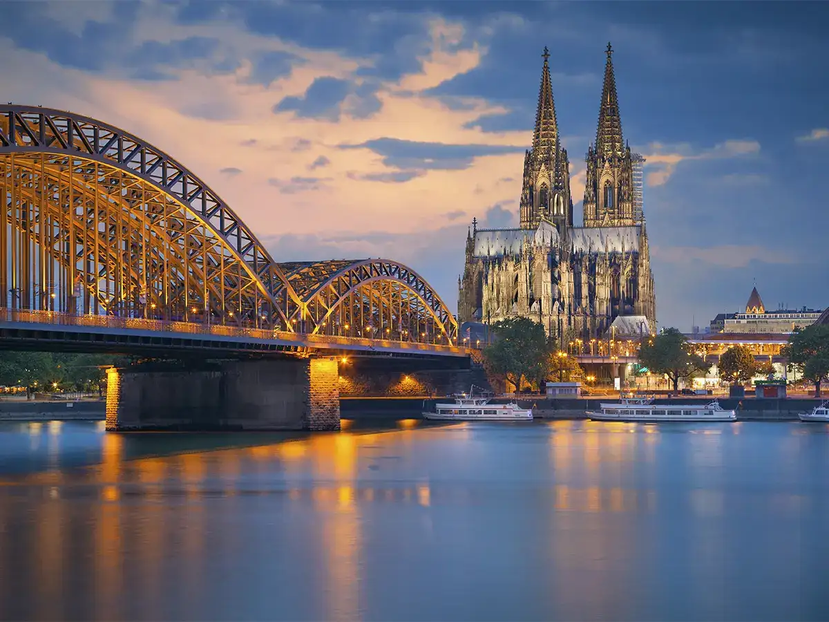 View of Cologne Cathedral and Hohenzollern Bridge in Cologne, Germany, with city lights reflecting on the Rhine River during twilight.