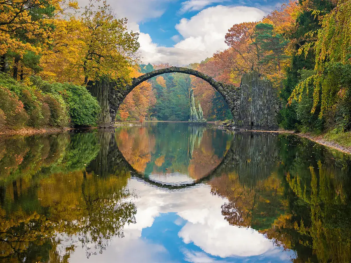 Ancient stone bridge over a calm river, surrounded by autumn trees. The bridge’s reflection creates a full circle with the water, under a partly cloudy sky.
