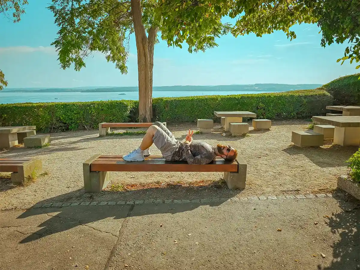 Relaxing in Birnau A person lies on their back on a bench in a park, with trees, picnic tables, and a body of water in the background.