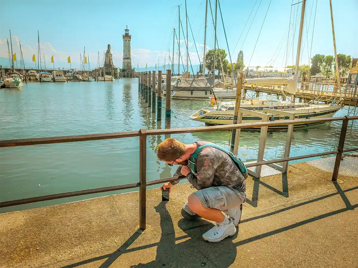 Catching boats in Lindau. A person kneels on a harbor walkway taking a photo with a smartphone; sailboats and a lighthouse are visible across the water on a sunny day.