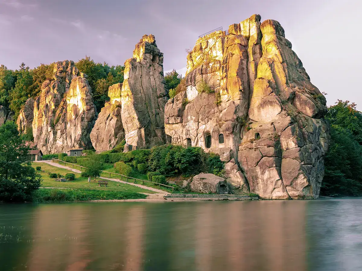 Rock formations rise near a calm body of water, with greenery surrounding them, under a softly lit sky.