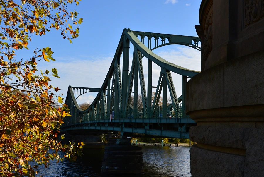 The green metal Glienicke  Bridge spans across a river. Autumn leaves frame the left side, and part of a stone building is visible on the right.