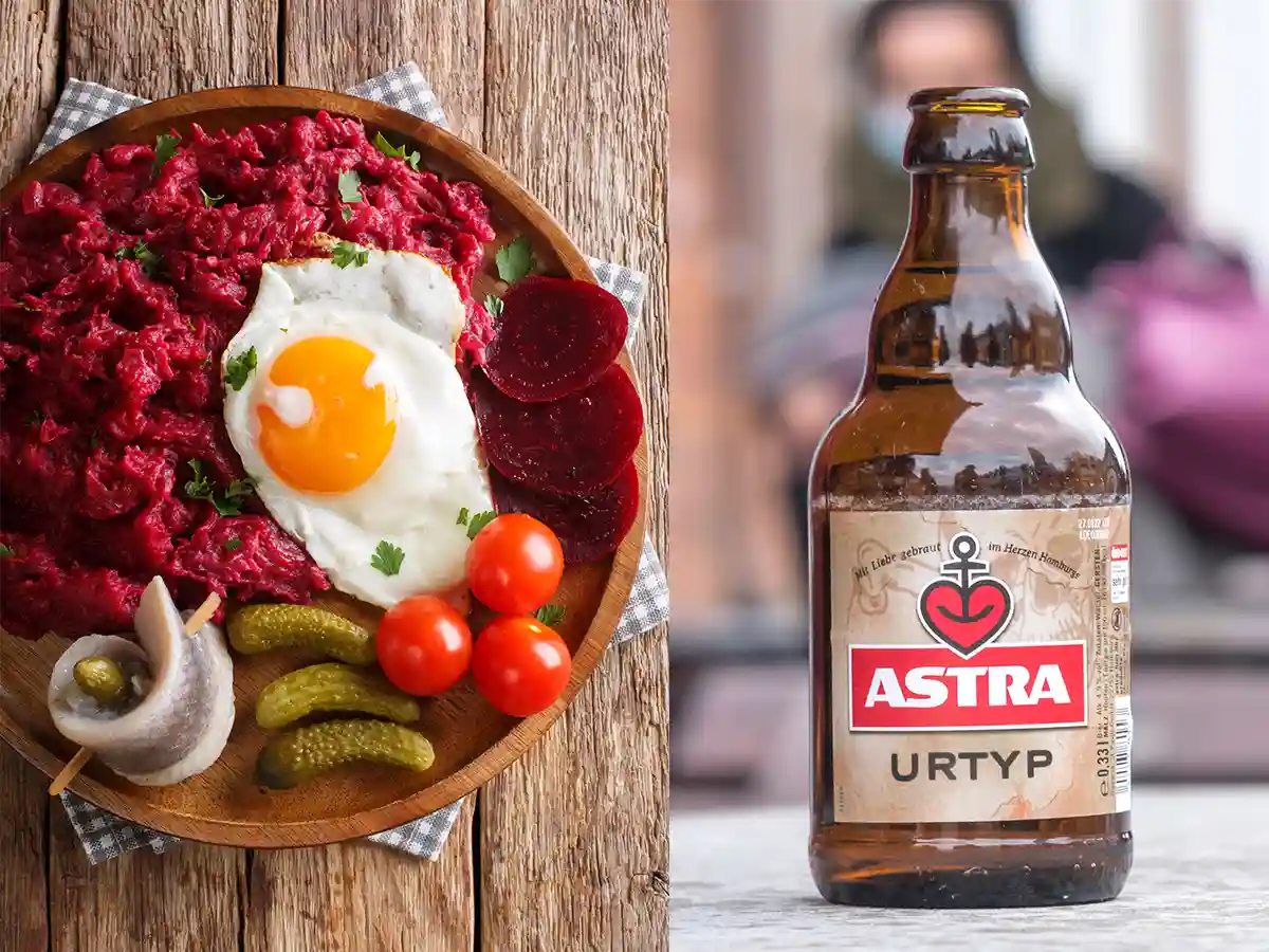 A wooden plate with Labskaus, topped with a fried egg, served with pickles, herring, tomatoes, and beetroot, next to a bottle of Astra Urtyp beer.