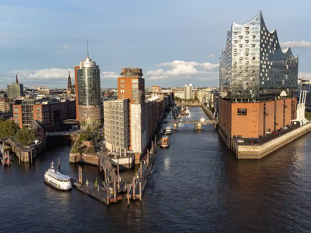 A riverside cityscape of Hamburg featuring modern and historic buildings along the water, with boats docked and the Elbphilharmonie concert hall visible on the right. A riverside cityscape of Hamburg featuring modern and historic buildings along the water, with boats docked and the Elbphilharmonie concert hall visible on the right.