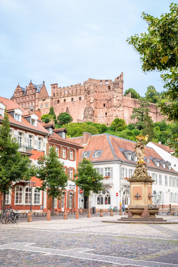 The Heidelberg Castle ruins on a hill overlook a town square with historic buildings and a statue-topped fountain surrounded by trees.