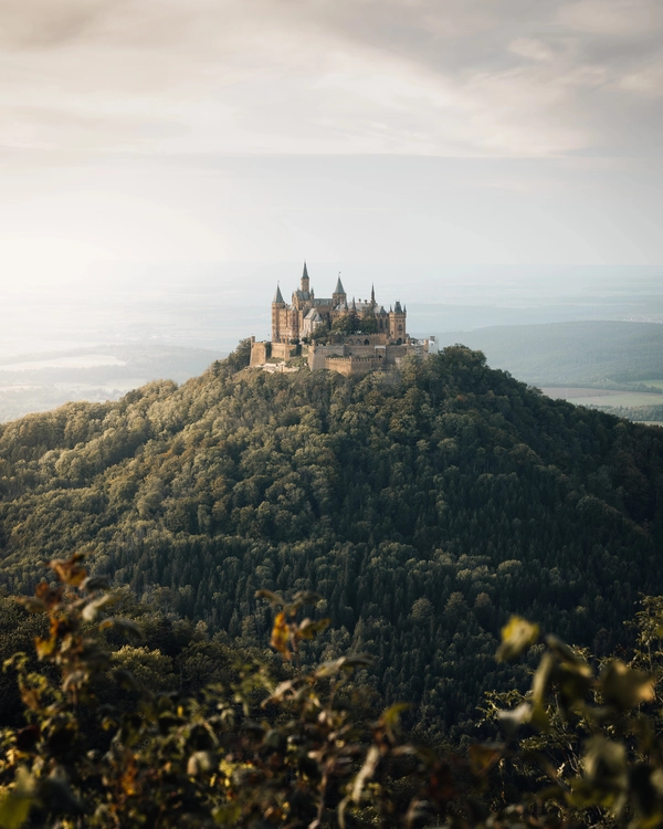 Hohenzollern Castle sits atop a forested hill under a cloudy sky, surrounded by mist and distant landscape scenery.