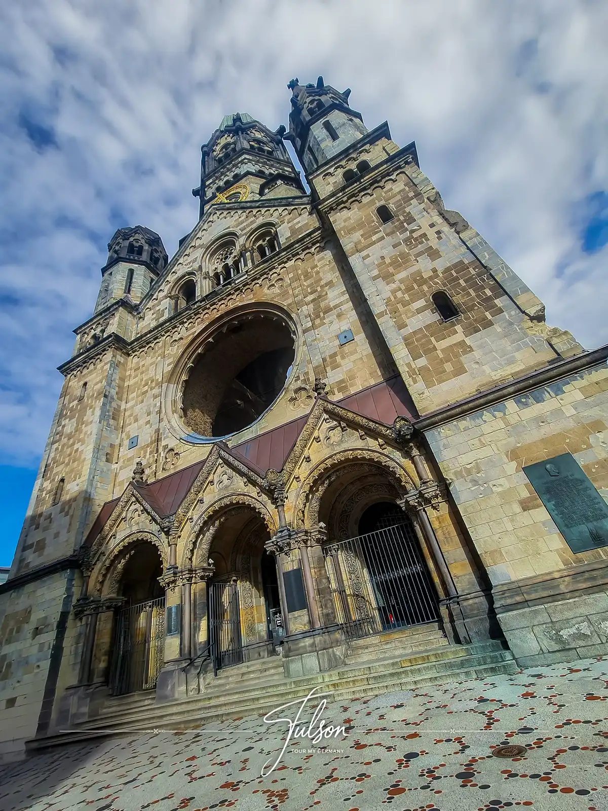 Low-angle view of the Kaiser Wilhelm Memorial Church in Berlin, showing its stone facade, arched entrances, and neo-Romanesque architecture against a partly cloudy sky.