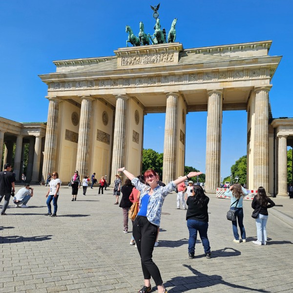 I mean, how could you miss it? Lydia, with outstretched arms, stands in front of the Brandenburg Gate in Berlin, with tourists and a bright blue sky in the background.