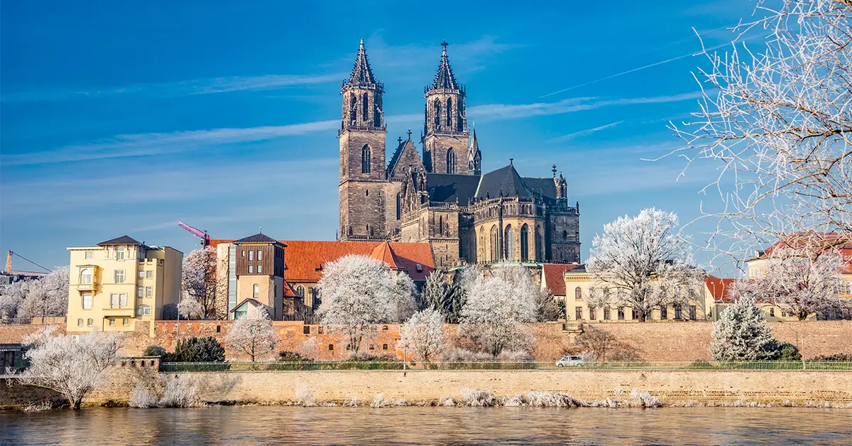 A historic cathedral in Magdeburg with twin towers stands surrounded by frosty trees and buildings, viewed from across a river under a clear blue sky.