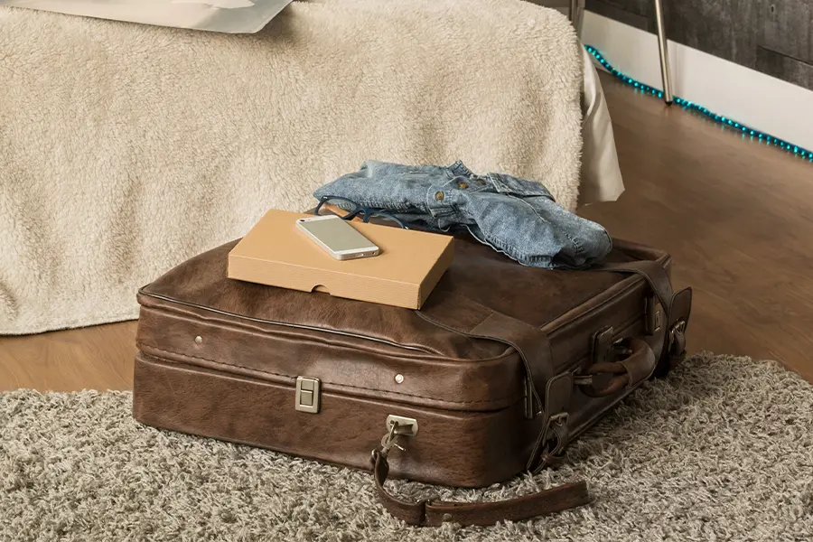 A brown leather suitcase on a rug, topped with a closed cardboard laptop box, a smartphone, and a folded denim shirt, positioned near a bed.
