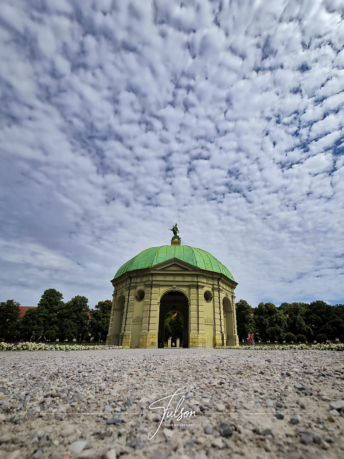 A green-domed pavilion in the English Garden in Munich stands under a cloudy sky, surrounded by trees and a gravel path in the foreground.