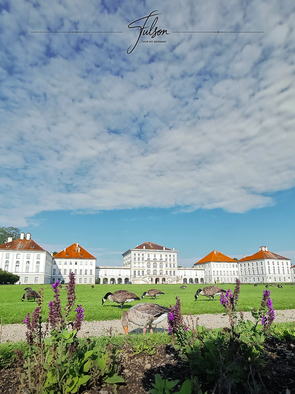 Geese grazing on the lawn in front of a large, symmetrical Nymphenburg Palace in Munich with a cloudy blue sky above.
