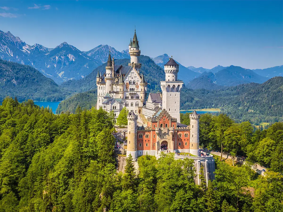 Aerial view of Neuschwanstein Castle in Germany, surrounded by lush green forests and distant mountains under a clear blue sky.