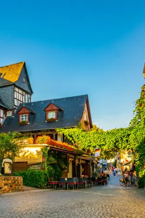 A charming street scene features a half-timbered house with ivy, outdoor café seating, and people walking on a cobblestone road under a clear blue sky.