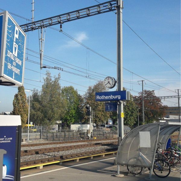 Rothenburg Train Station Train station platform in Rothenburg with signage, overhead wires, and a clock reading the time. Bicycle parking area and trees are visible in the background.