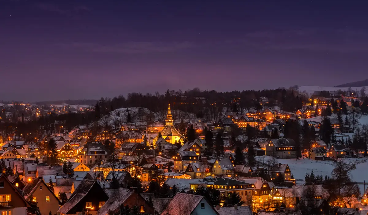 Snow-covered town at dusk with illuminated buildings and a prominent church steeple, surrounded by hills and trees under a clear sky.