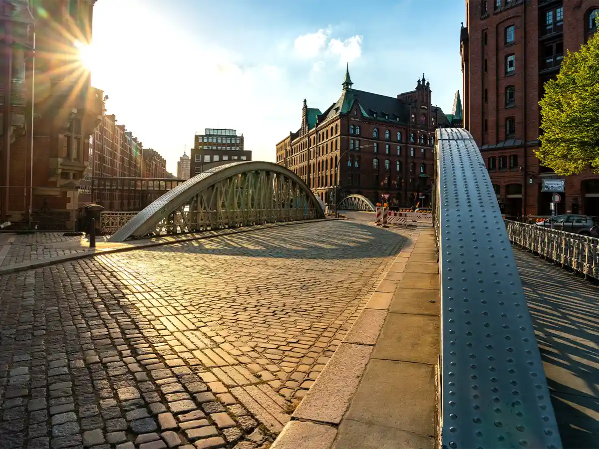 A cobblestone street leads to a metal arch bridge surrounded by historic brick buildings under a bright, partly cloudy sky. A cobblestone street leads to a metal arch bridge surrounded by historic brick buildings under a bright, partly cloudy sky.