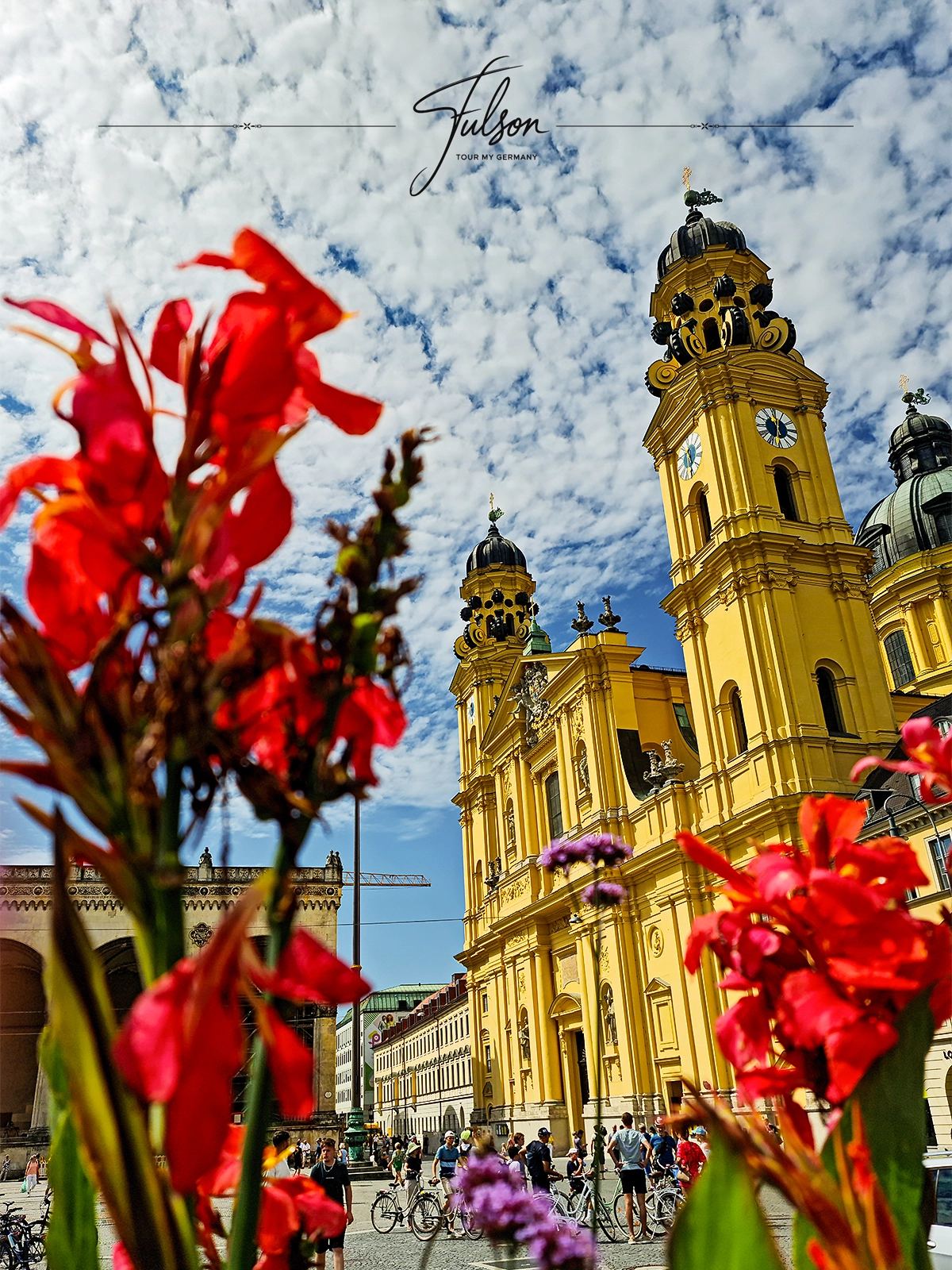 The vibrant yellow Theatinerkirche in Munich with ornate towers stands under a partly cloudy sky, framed by red flowers. People and bicycles are visible in the plaza below.