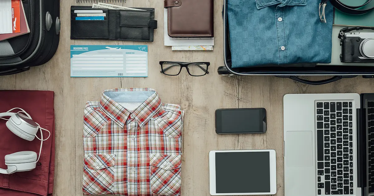Organized travel essentials on a table: open suitcase with clothes, camera, laptop, tablet, smartphone, headphones, glasses, wallet, boarding pass, and travel documents.