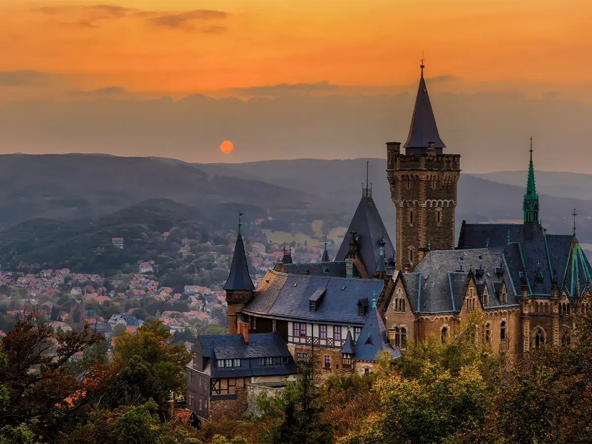 Castle with multiple towers in a hilly landscape at sunset. The sky is orange, and a sun sets behind distant hills. Trees and a small town are visible in the background.