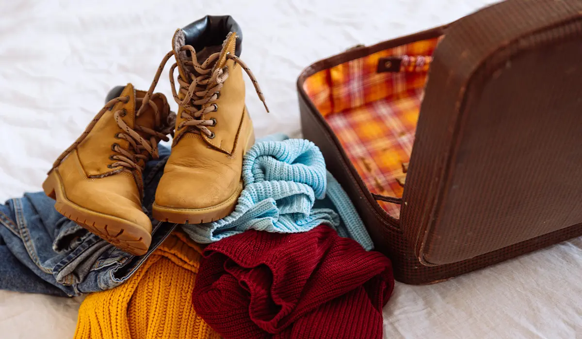 Brown boots, folded sweaters in red, yellow, and blue, and jeans are placed beside an open, empty suitcase on a bed with a white sheet.