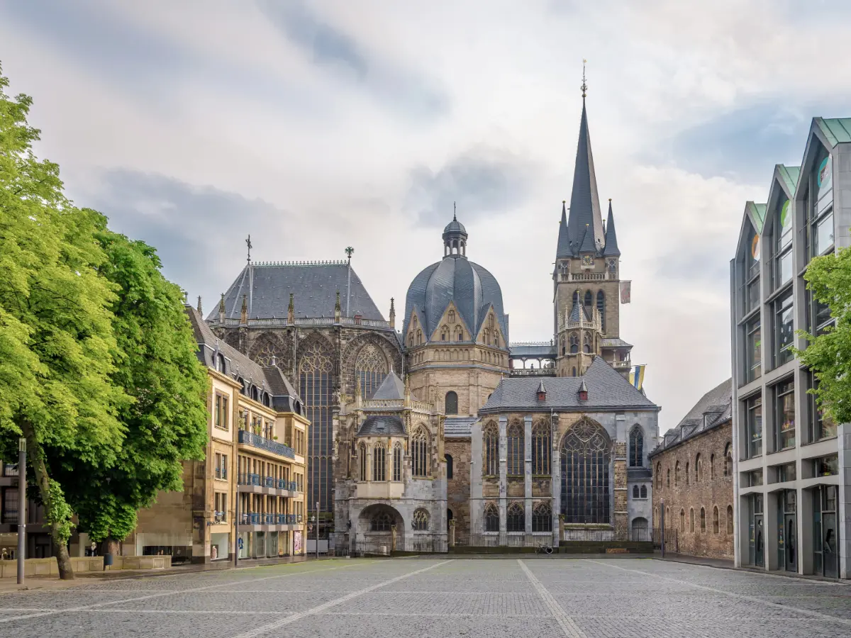 Aachen Cathedral in Germany, featuring a mixture of architectural styles with its prominent dome and spires, stands surrounded by historic buildings and a cobblestone plaza.