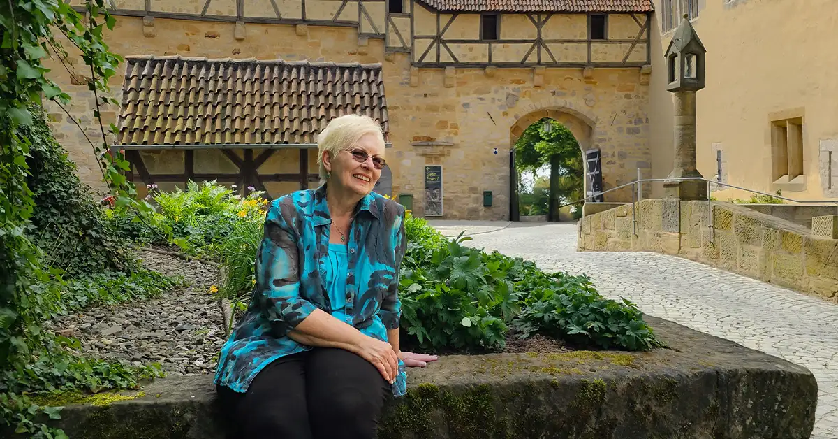 Woman sitting on a stone ledge in front of a medieval building with timber framing and a cobblestone path, smiling on a sunny day.