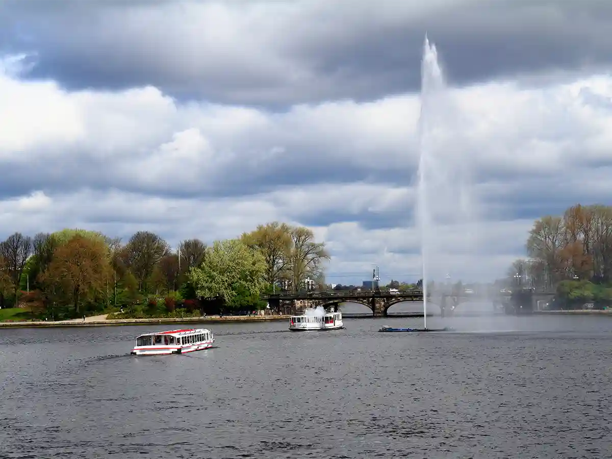 Two white boats on Alster Lakes in Hamburg near a tall water fountain, with a bridge and trees in the background under a cloudy sky. Two white boats on Alster Lakes in Hamburg near a tall water fountain, with a bridge and trees in the background under a cloudy sky.