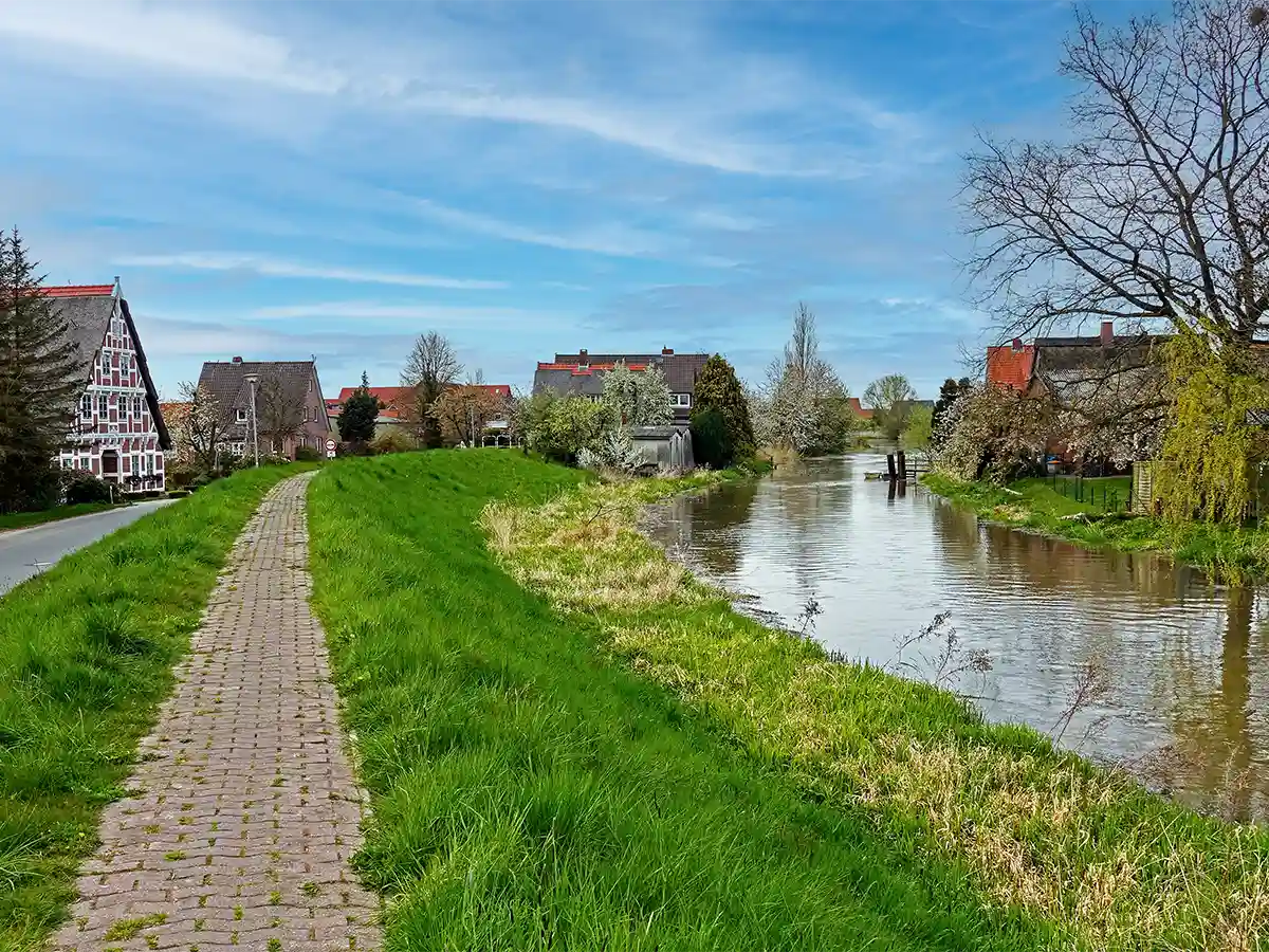 A paved walkway runs alongside a grassy embankment next to a calm river with houses and trees in the background under a blue sky. A paved walkway runs alongside a grassy embankment next to a calm river with houses and trees in the background under a blue sky.