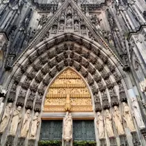 Gothic stone archway of Cologne Cathedral's main entrance, with rows of carved saints and golden reliefs above the doorway.