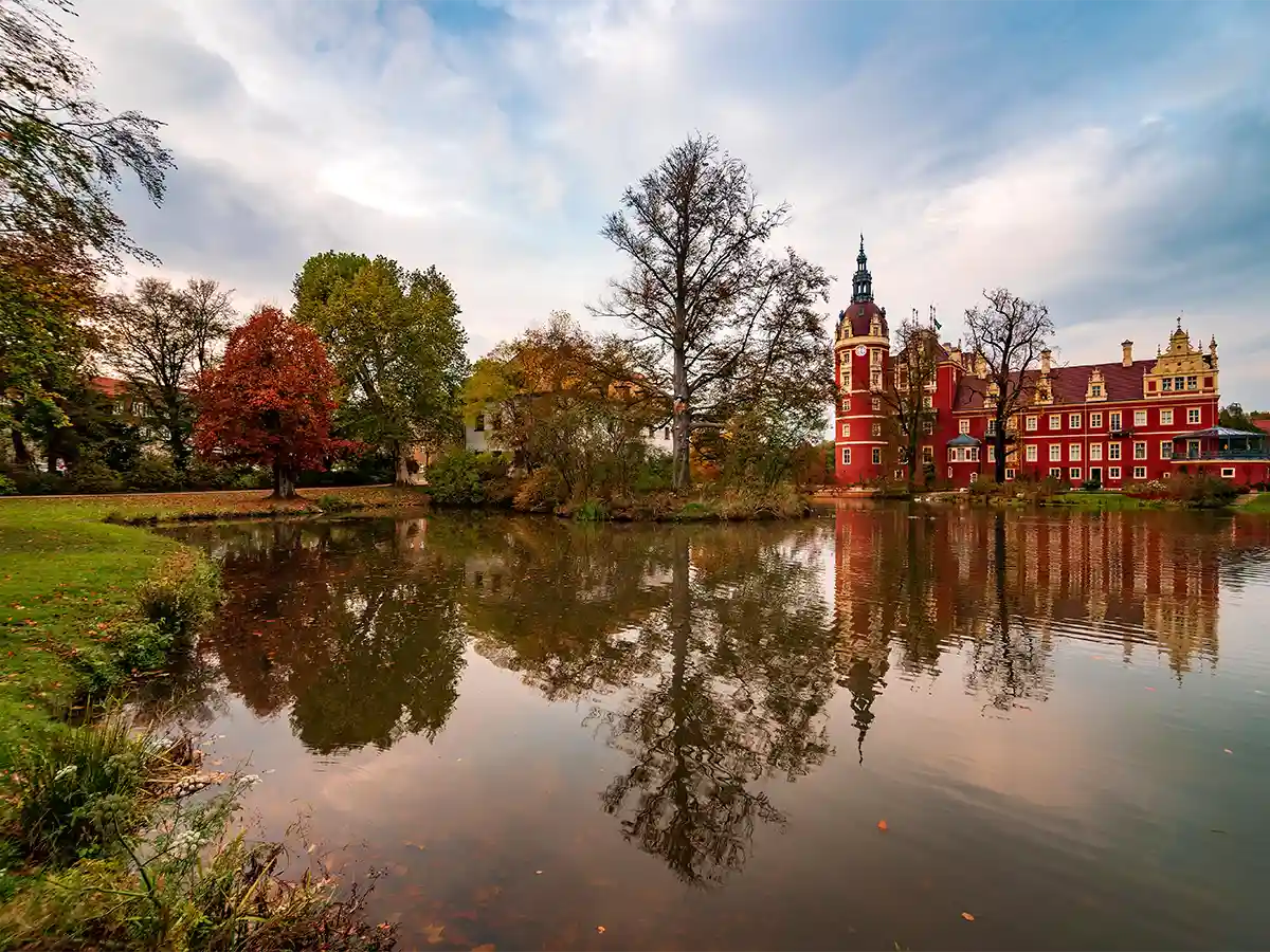 Muskau Park’s red Neues Schloss reflected in calm lake, autumn colours blazing—classic Brandenburg castles and lakes.