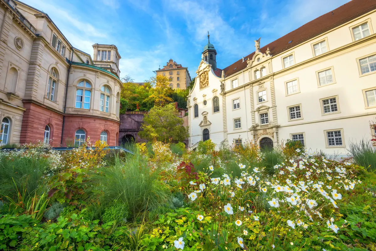 A scenic view of a historic European building complex in Baden-Baden with ornate architecture, surrounded by lush greenery and white flowers under a bright blue sky.