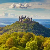 Hohenzollern Castle perched on a lush green hilltop, surrounded by trees and overlooking a vast landscape under a partly cloudy sky.