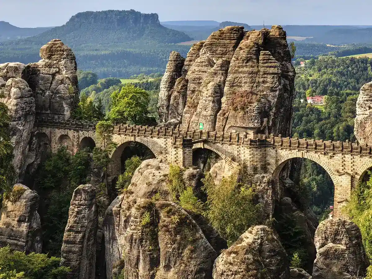 Stone bridge with arches spans between large rocky formations surrounded by greenery, with a distant forested mountain under a clear sky.