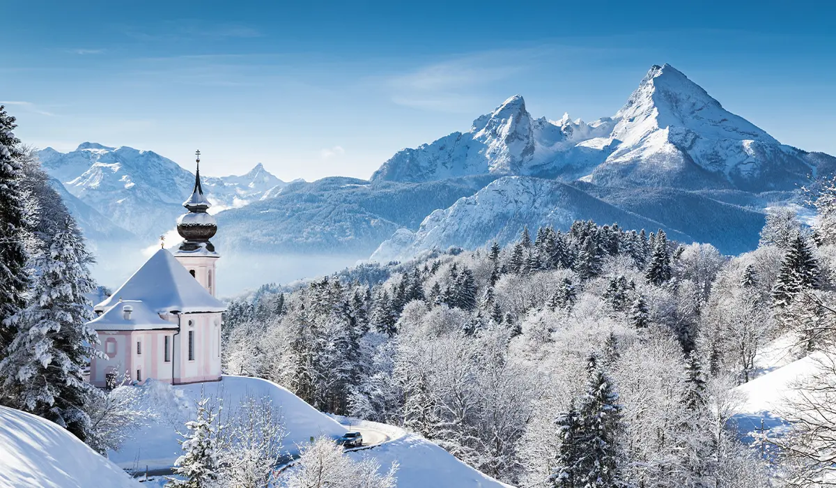 A snow-covered church on a hilltop with a backdrop of snowy mountains and a clear blue sky in the Bavarian Alps.