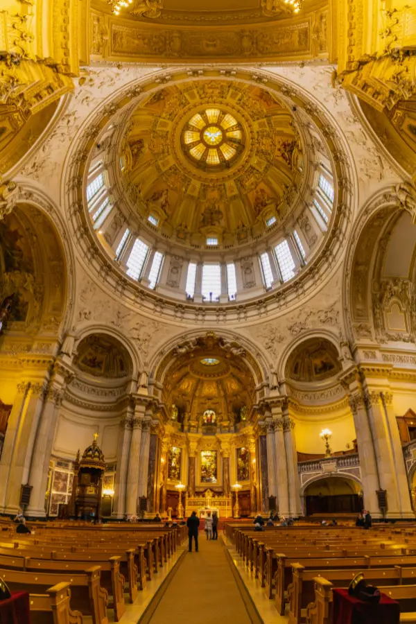 Interior view of Berlin Cathedral with a grand domed ceiling, intricate carvings, and rows of pews. The image captures the central nave and the altar area.
