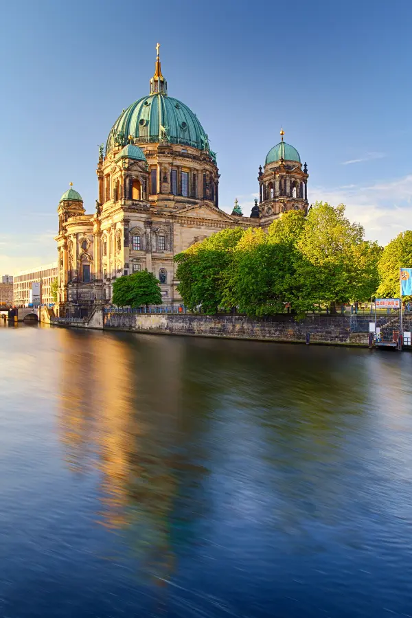 Berlin Cathedral: A large, historic building with a green dome is reflected in a calm river; trees and clear sky surround it.