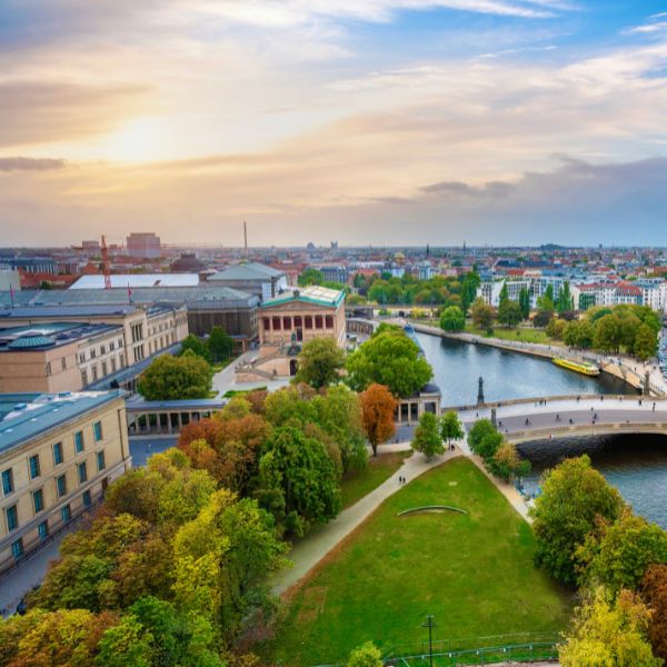 Museum Island, Berlin. On the left. Aerial view of a cityscape showing a river flanked by buildings and green spaces with a bridge connecting the riverbanks under a blue, cloudy sky at sunset.