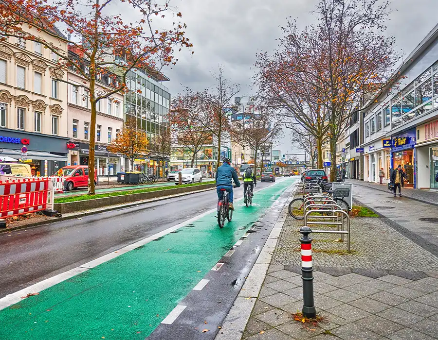 Cyclists ride along a green bike lane through central Berlin on a gray November day, surrounded by shops, trees, and passing cars.