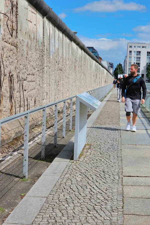 A person walks along a paved path beside a section of the Berlin Wall, under a bright blue sky.