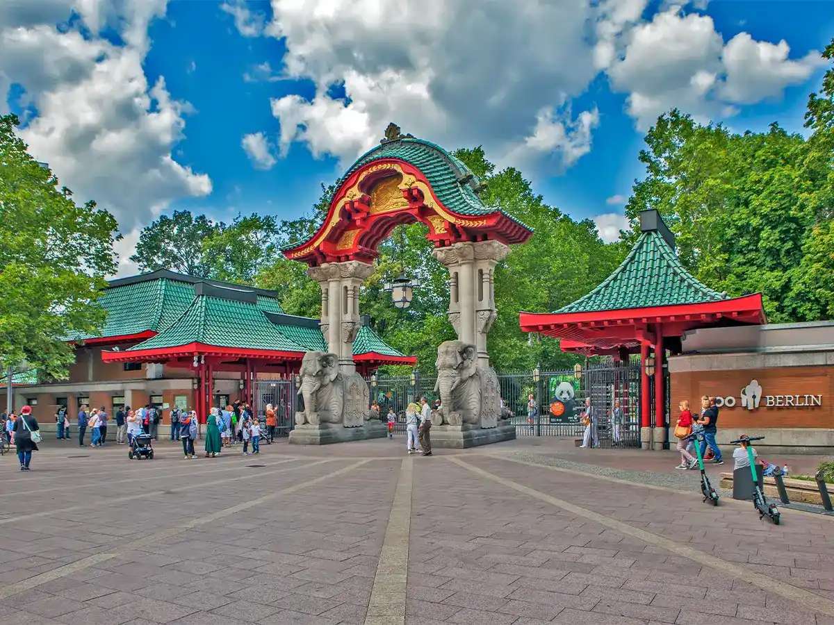 Visitors gather at the ornate entrance of Berlin Zoo, featuring two stone elephants and red, green-roofed structures, under a partly cloudy sky.