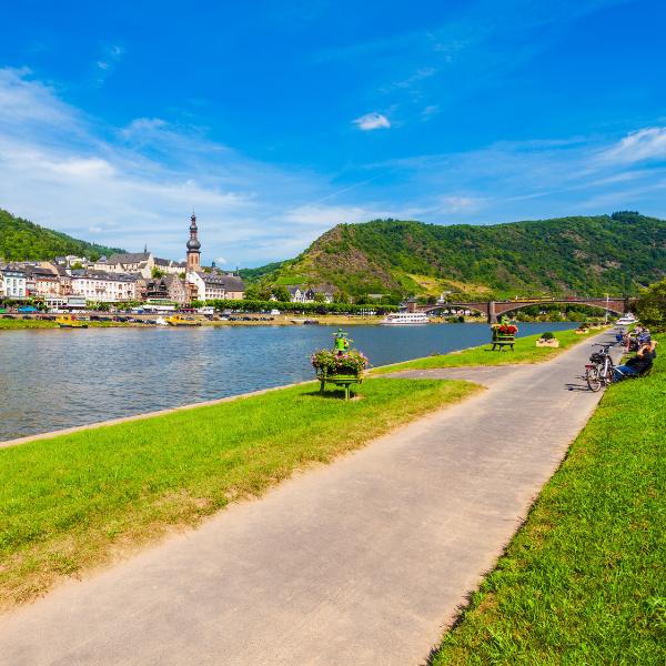 These paved paths go on for miles along the Mosel River. Enjoy the view! A paved riverside path beside a grassy area with benches and people on bicycles. The path overlooks a river, with a town and green hills in the background under a clear blue sky.